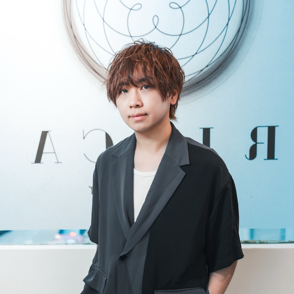 A handsome japanese style top hair stylist  with short brown hair stands wearing a black asymmetrical blazer over a white shirt and dark gray trousers. The background is a clean white wall with a circular graphic and stylized lettering. 